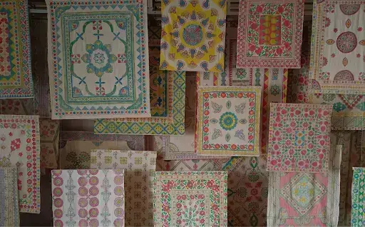 Colorful, patterned textiles hang vertically, featuring intricate floral and geometric designs in teal, pink, yellow, and green.