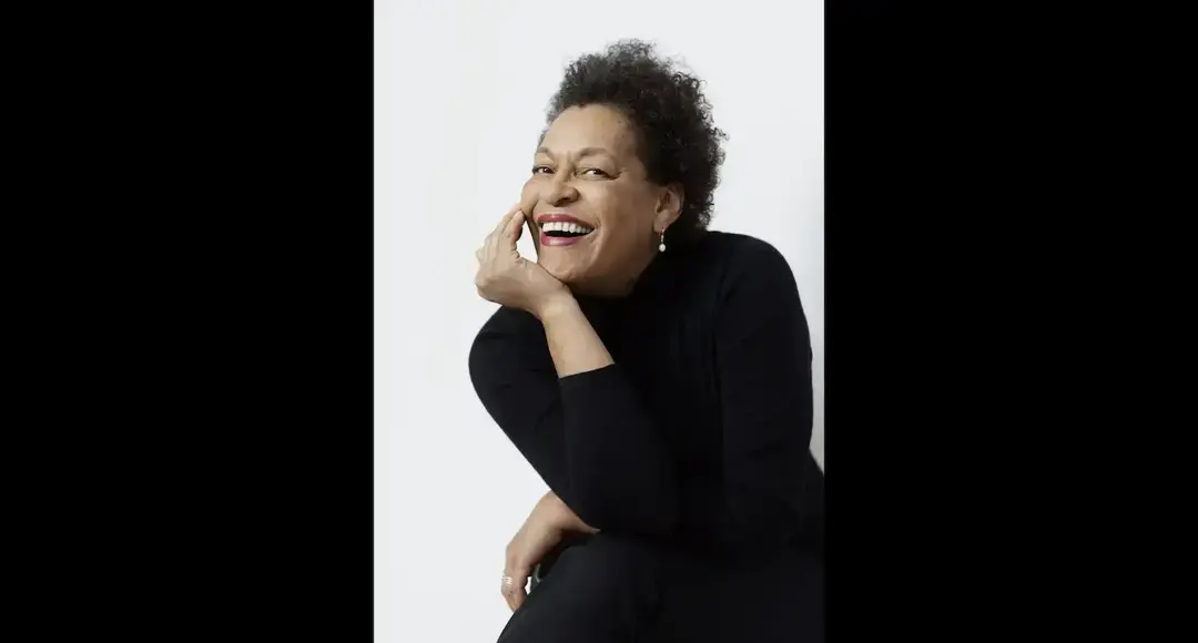 A smiling woman with short curly hair and pearl earrings in a black turtleneck rests her chin on her hand.