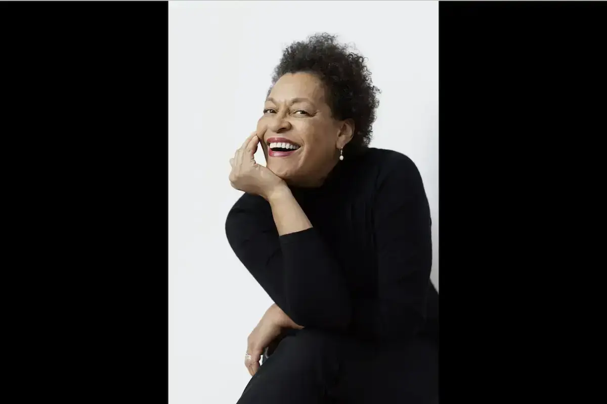 A smiling woman with short curly hair and pearl earrings in a black turtleneck rests her chin on her hand.