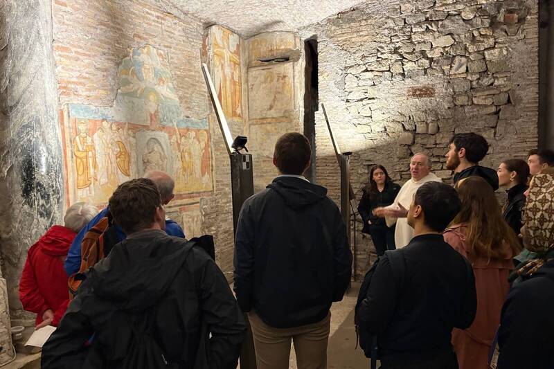 A priest in white robes gestures while guiding a diverse group of visitors past ancient, colorful frescos on brick walls.