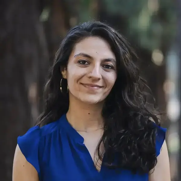 Smiling woman with dark wavy hair and gold hoop earrings wears a royal blue v-neck top outdoors.