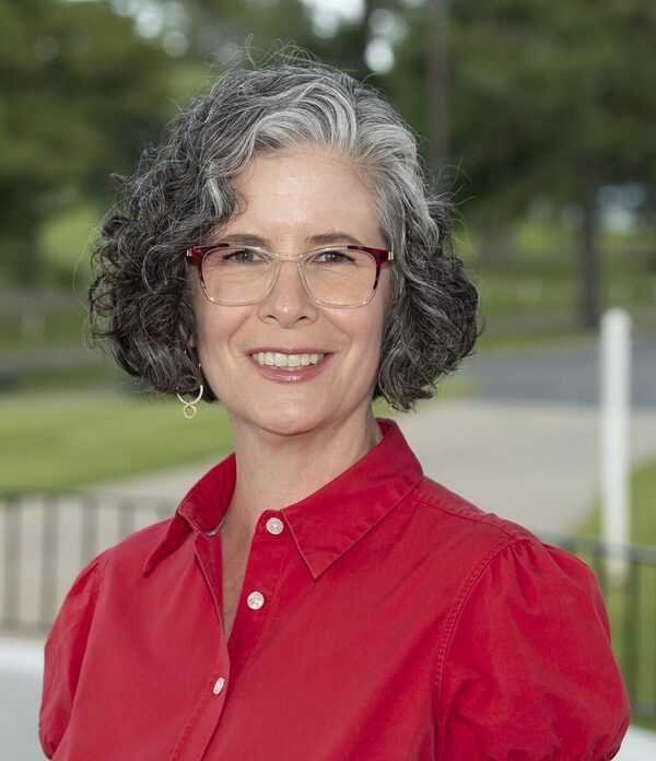 Smiling woman with short, curly gray hair and red-framed glasses. She wears a red collared shirt against a blurred green background.