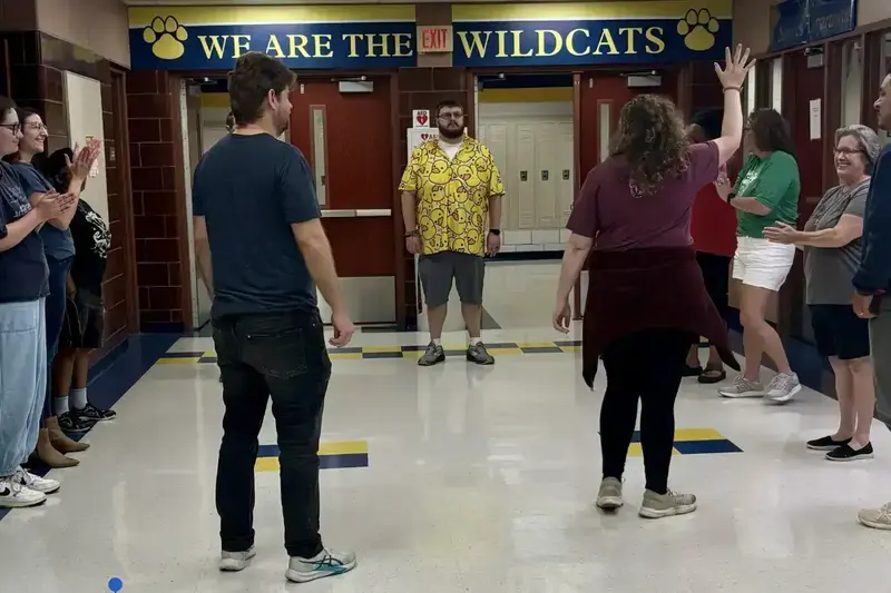 Man in yellow duck shirt welcomed by a line of clapping, smiling people in a Wildcats school hallway.