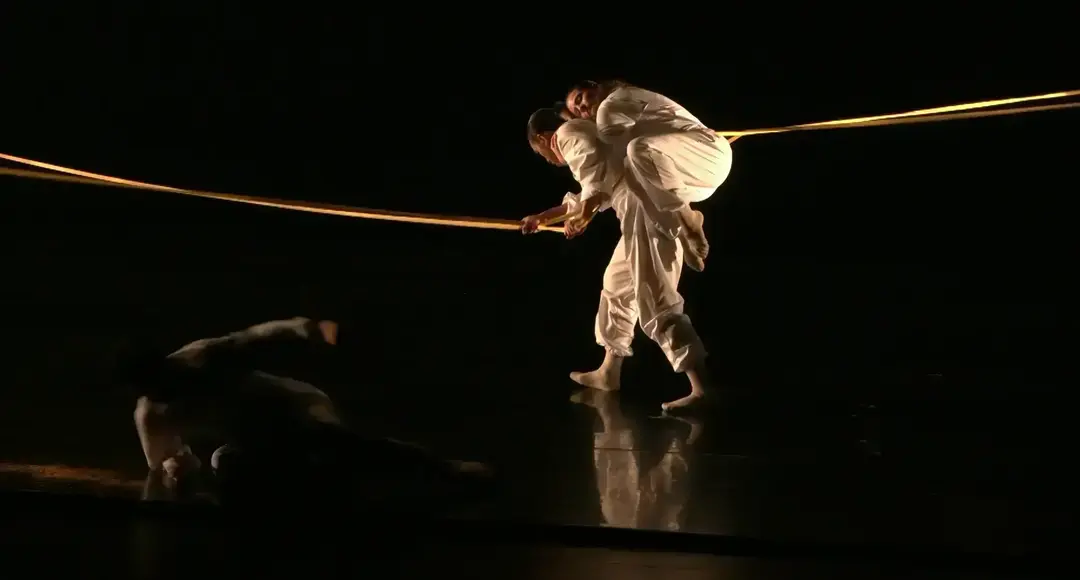 Two barefoot dancers in white, one carrying the other, hold a glowing golden rope on a dark stage. A third dancer is on the floor.