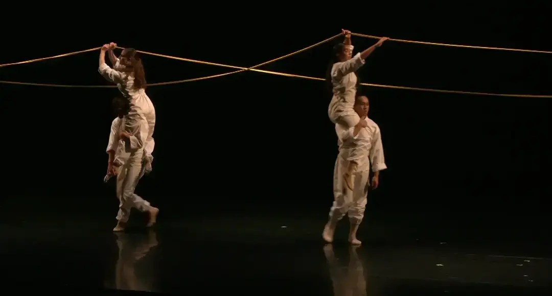 Two pairs of dancers in white balance on shoulders, holding golden ropes that crisscross on a dark stage.
