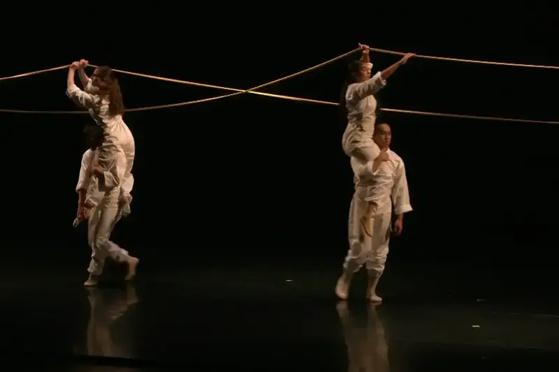 Two pairs of dancers in white balance on shoulders, holding golden ropes that crisscross on a dark stage.